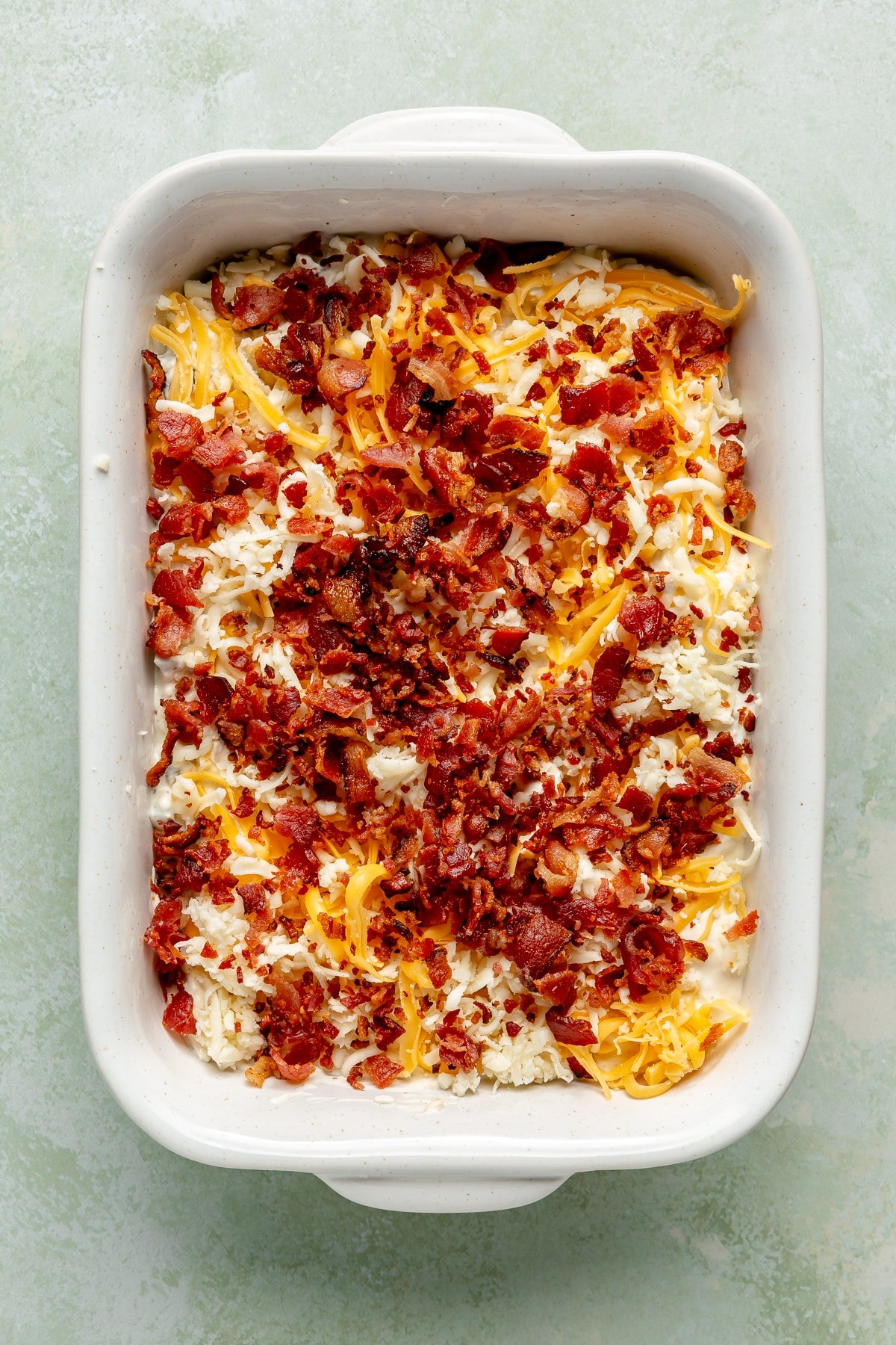 The cream cheese mixture has been placed into a white casserole dish. It has been topped with shredded cheese and bacon bits.