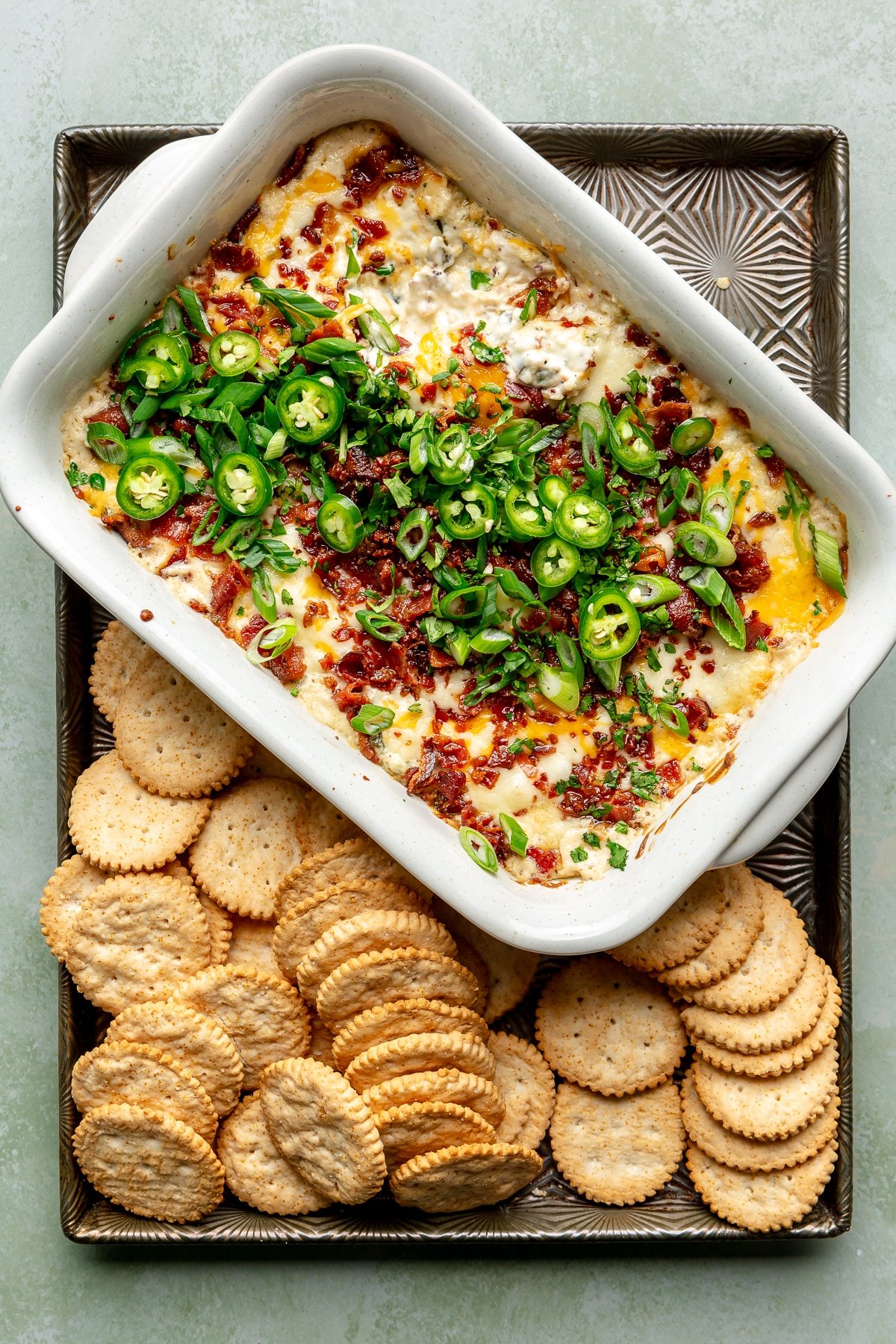 Jalapeno popper dip, which still sits in its casserole dish, has been topped with diced jalapeños. It sits on a tray alongside round crackers.