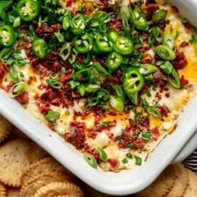 Jalapeno popper dip, which still sits in its casserole dish, has been topped with diced jalapeños. It sits on a tray alongside round crackers.