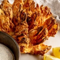 An air fryer blooming onion sits on a plate next to a bowl of sauce and lemon wedges.