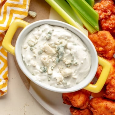 Blue cheese dip has been placed into a white serving dish with yellow handles. It has been topped with blue cheese crumbled. It sits next to pieces of breaded chicken and celery sticks.