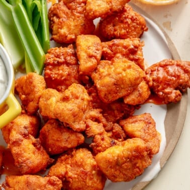 Buffalo chicken bites sit on a serving plate next to a side of blue cheese dip. Pieces of celery sit to the side.