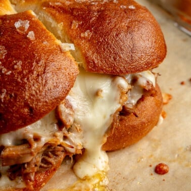 Fully prepared pulled pork sliders sit on a baking sheet. Melted cheese is shown dripping down the side.