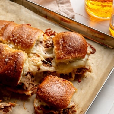 Fully prepared pulled pork sliders sit on a baking sheet. Two sliders are shown being pulled away from each other to show the cheese pull.