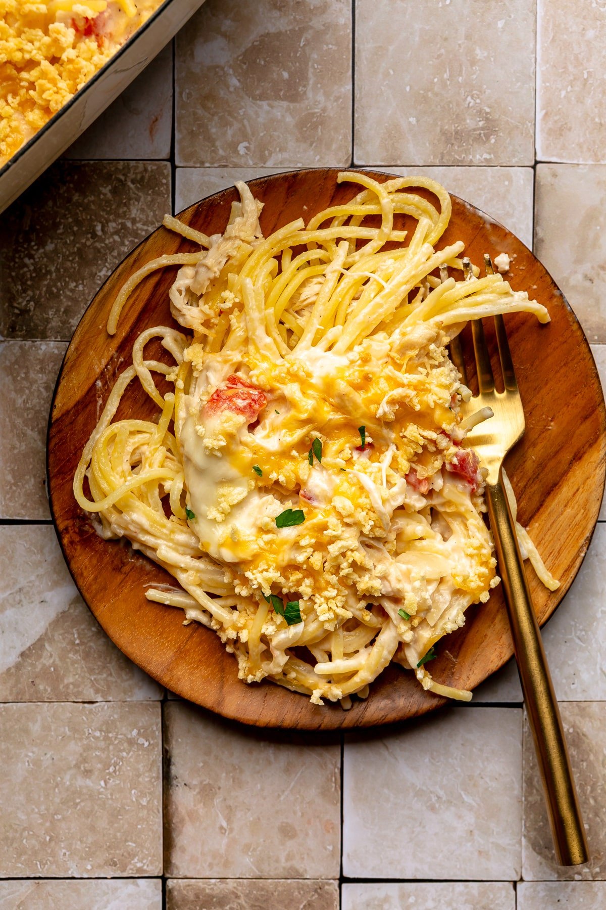 Spaghetti, topped with the baked chicken mixture, sits on a wooden serving plate. A gold colored spoon sits to the side.