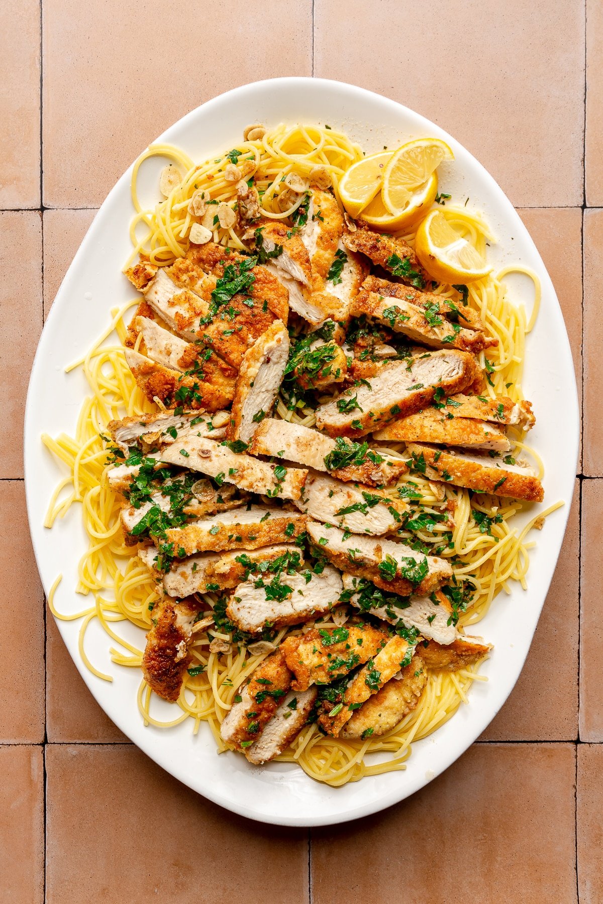 Chicken scampi sits on a bed of spaghetti on a white serving plate. Slices of lemon sit to the side.