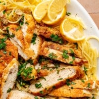 Chicken scampi sits on a bed of spaghetti on a white serving plate. Slices of lemon sit to the side.