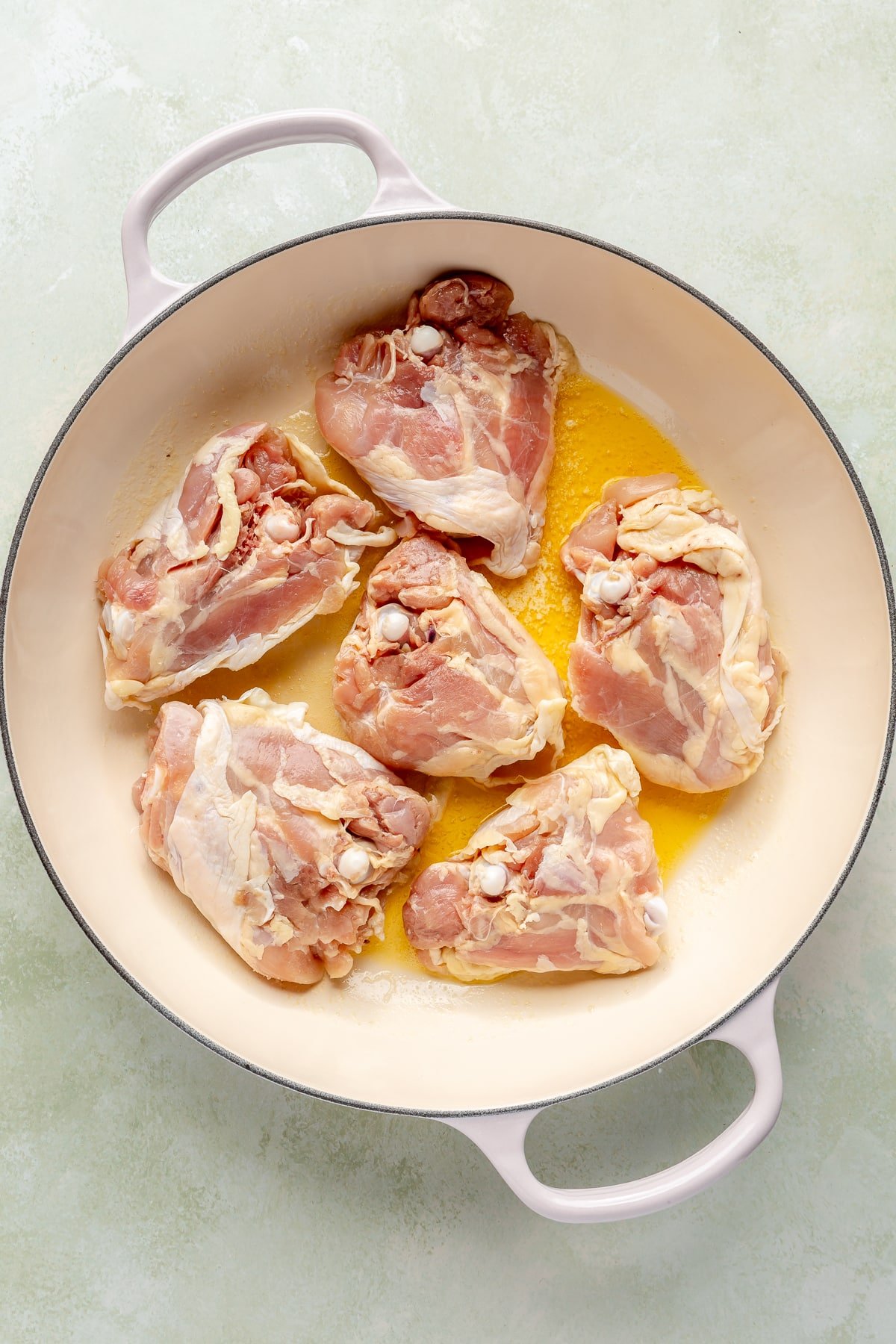 6 chicken thighs have been placed in a white enameled pot with a yellow oil.