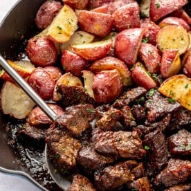 Fully prepared garlic butter steak and potatoes sit in a cast iron skillet. A metal serving spoon is shown scooping up some of the steak.