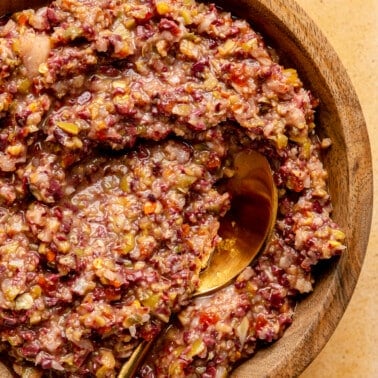 Olive tapenade sits in a wooden serving bowl. A gold colored spoon sits to the side.