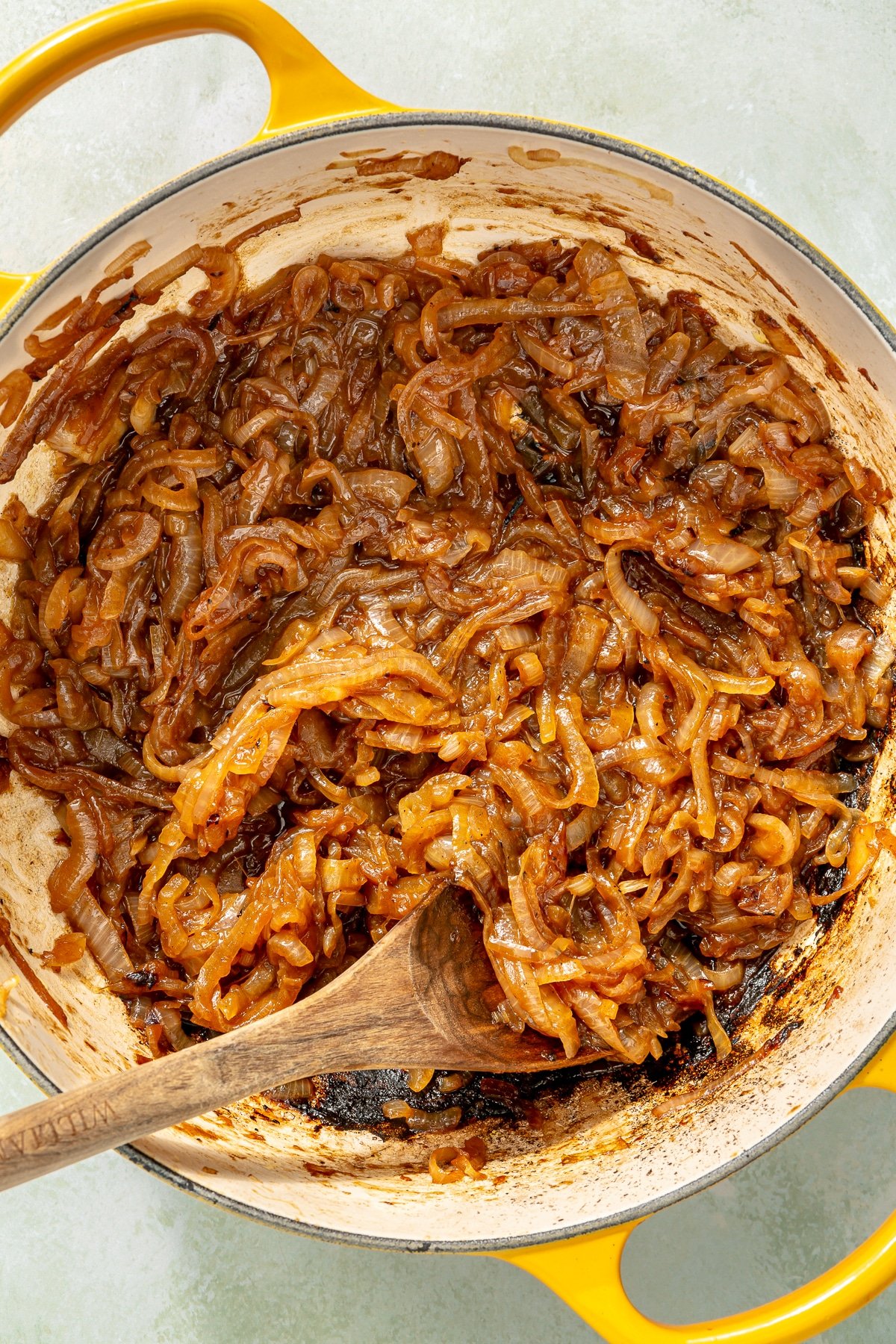 Caramelized onions sit in an enameled pot with yellow handles. A wooden spoon is shown mixing the onions.