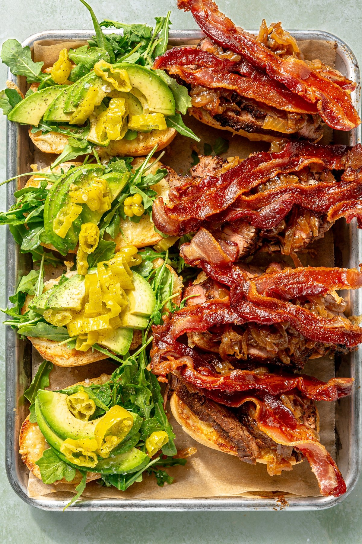 Sliced steak, bacon, caramelized onions, avocado slices, and pepperocinis have been placed on the toasted baguette halves.