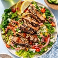 Fully prepared balsamic steak salad sits in a white bowl on a light blue surface. A glass of ice water, as well as a bowl of halved cherry tomatoes and a bowl of sliced cucumbers, sits to the side.