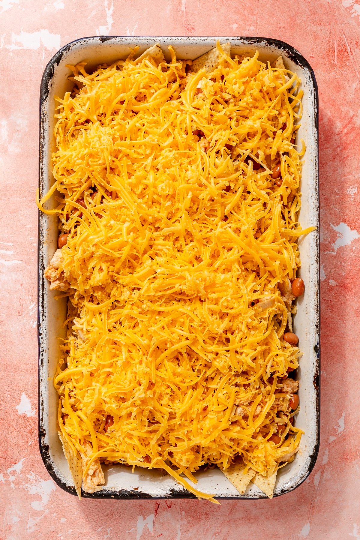 Fully assembled nachos sit on a white sheet pan. Shredded cheddar cheese covers them.