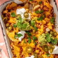 A hand is shown lifting a nacho from the pan. Dollops of sour cream and guacamole sit on top.
