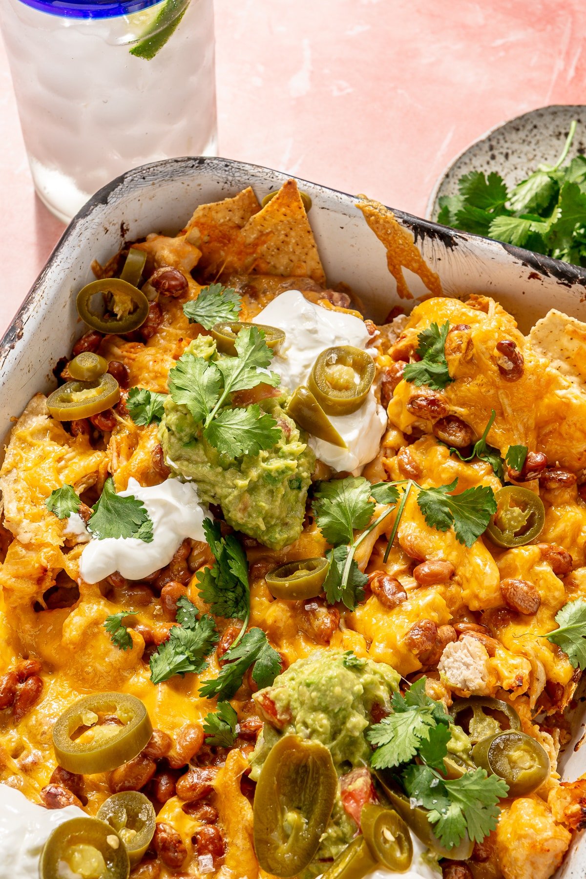 Sheet pan nachos sit in a white colored sheet pan. A glass of ice water with a slice of limes sits to the side.