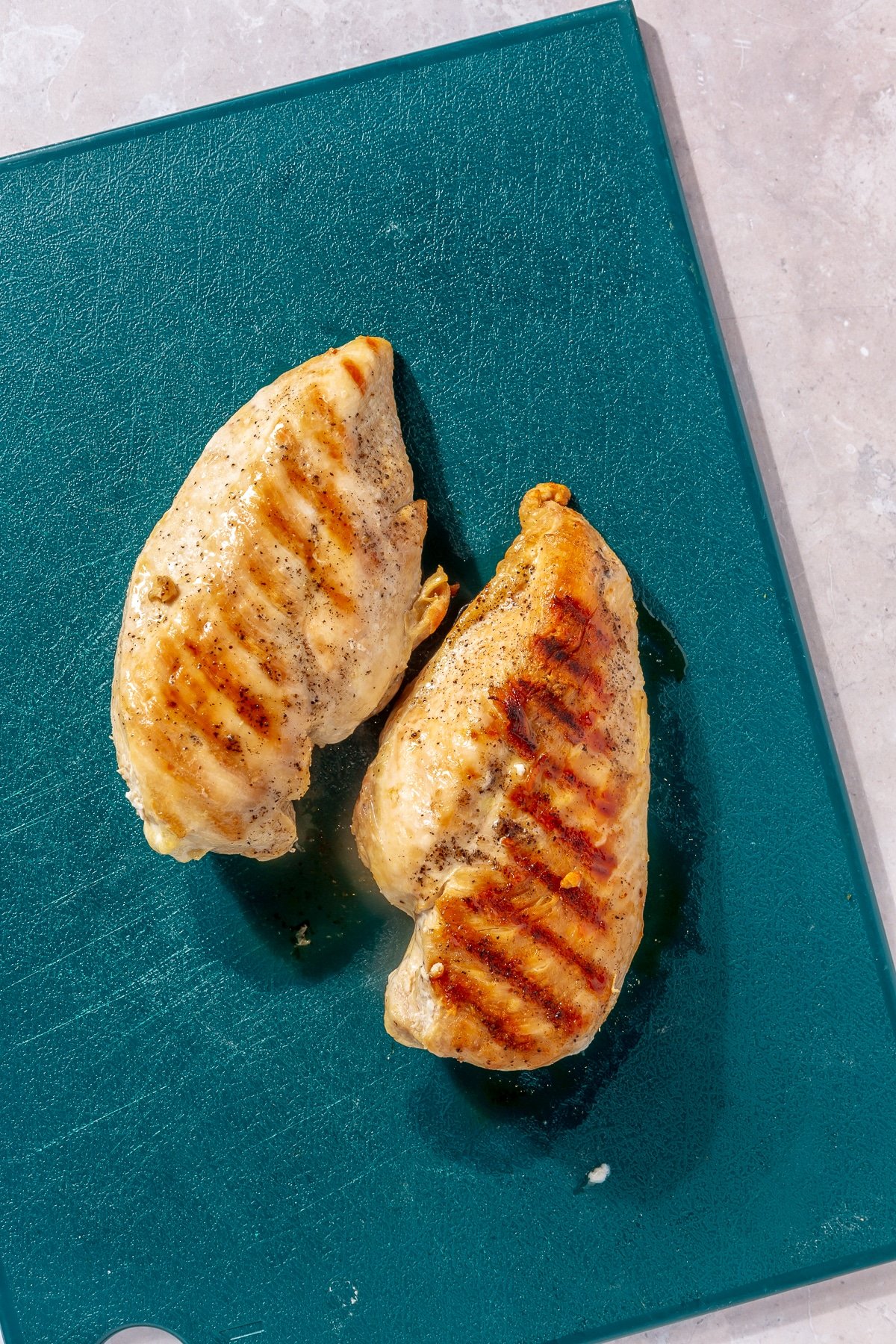 Two grilled chicken breasts sit on a teal colored cutting board.