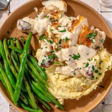 Slices of chicken breast sit on a brown colored plate next to a side of mashed potatoes and green beans.