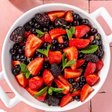 Mixed berry salad sits in a white serving bowl on a pink tiled surface.