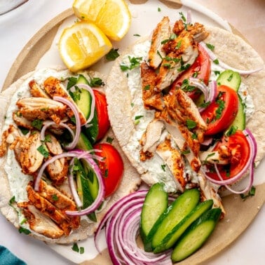 Two chicken gyros sit on a white and brown colored plate. A plate of tomato slices sits to the side.
