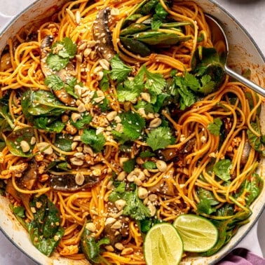 Fully prepared gochujang pasta sits in a large white pot. Chopped peanuts and slices of lime sit to the side.