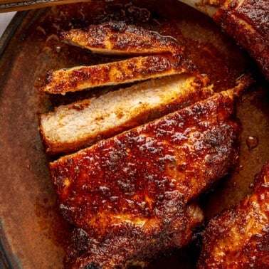 Three cooked and glazed pork chops sit on a brown colored plate. One has been sliced to show the inside.