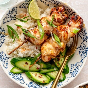 Two bang bang chicken skewers sit on a bed of white rice next to sliced cucumbers.