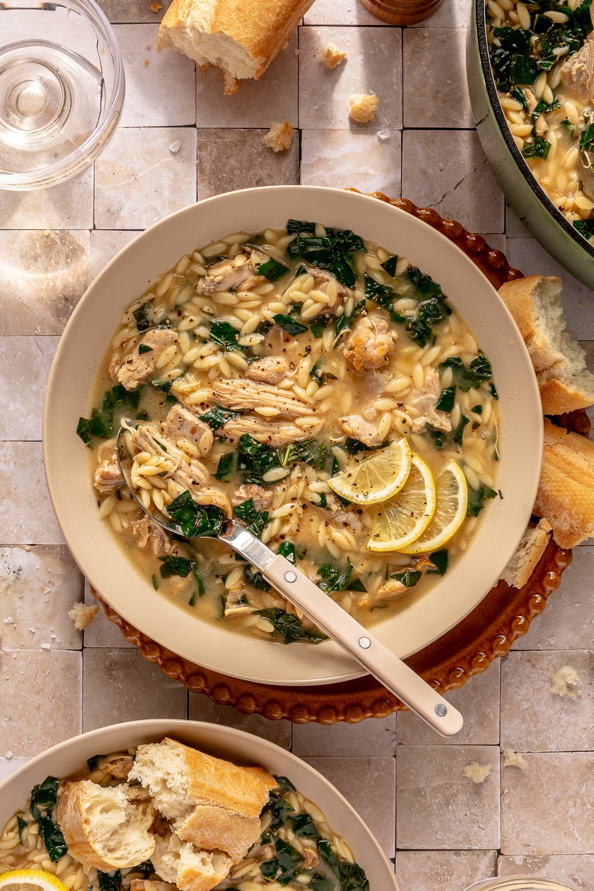 A bowl of Lemon Chicken Orzo soup ready to eat and surrounded by crusty bread and a glass of clear liquid.