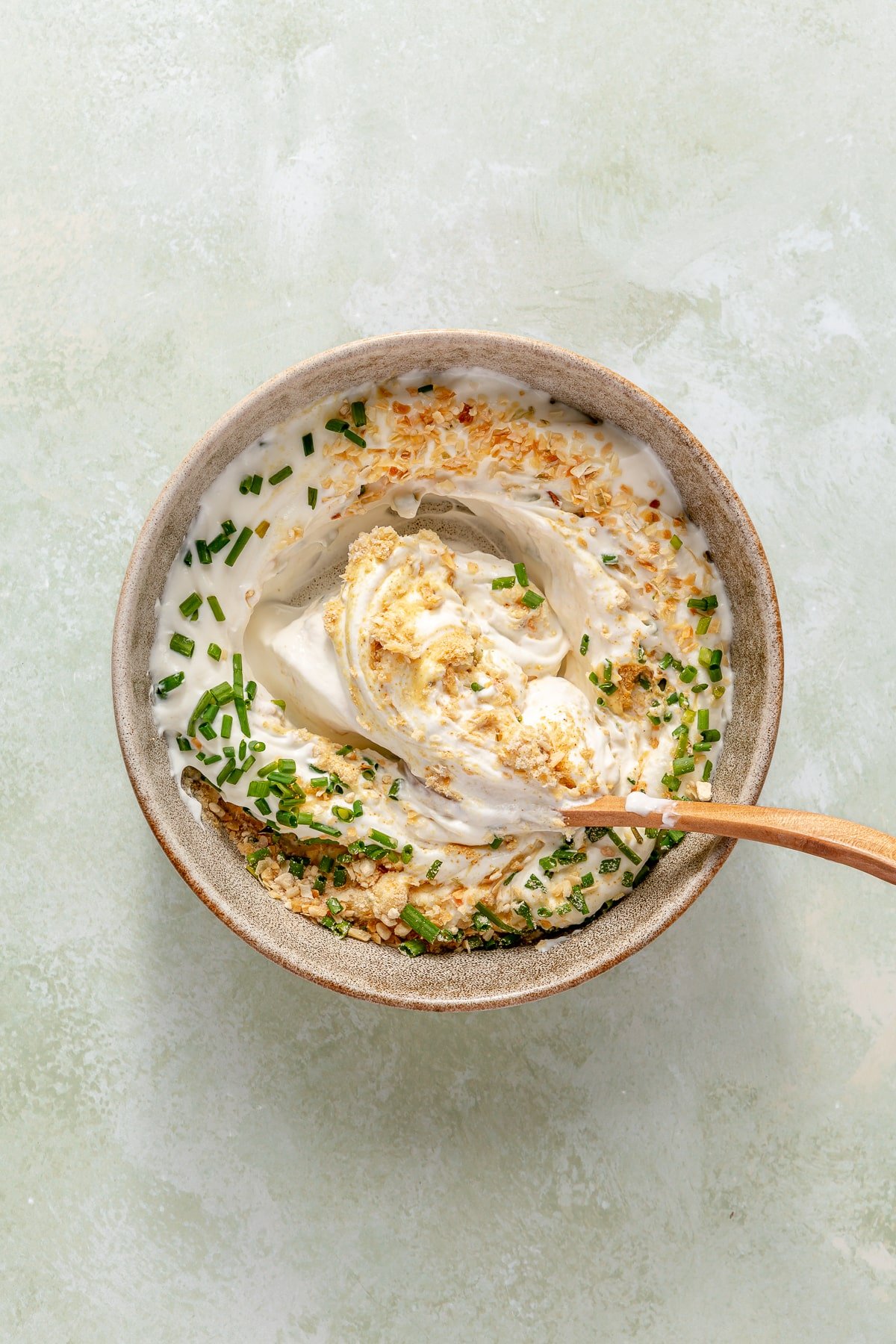 A bowl of Greek yogurt, sour cream, and all of the seasonings needed for homemade French onion dip, and a person using a spoon to stir the ingredients together.