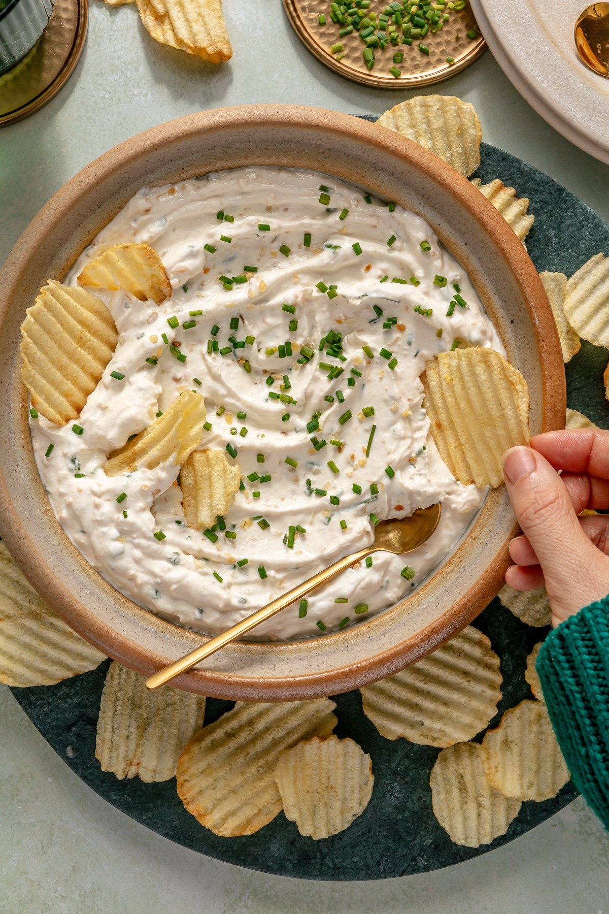 A large bowl of French onion dip garnished with fresh chives and surrounded by potato chips, and a person scooping some dip with a potato chip.