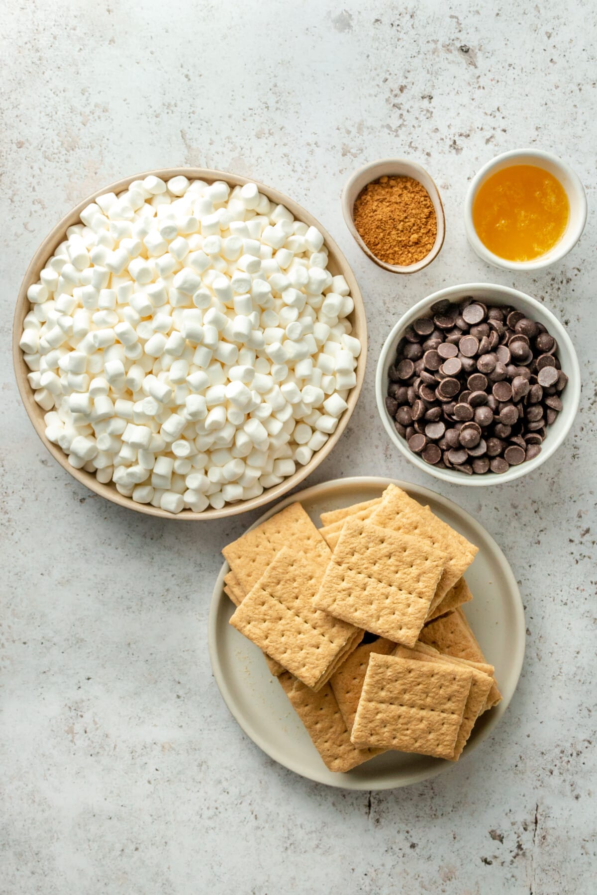 Ingredients for 5 ingredient smores bars sit in a variety of plates and bowls on a light grey surface.