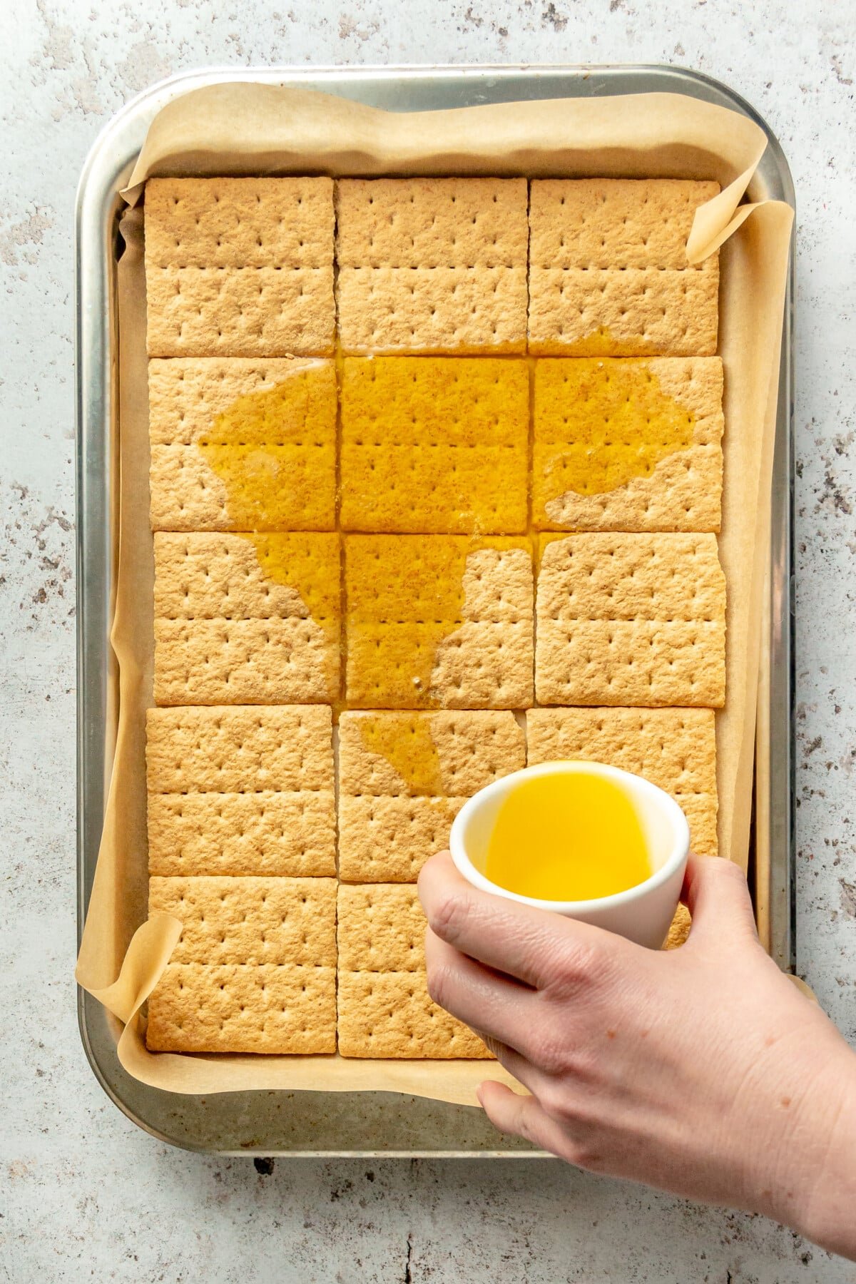 Melted butter is poured over graham crackers sitting on a lined and rimmed baking sheet on a light grey surface.