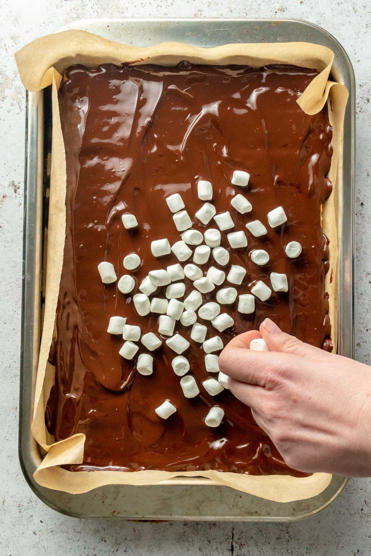 Marshmallows are scattered over melted chocolate on a lined rimmed baking sheet on a light grey surface.