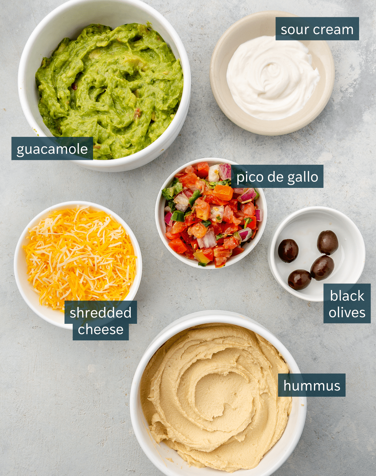 All of the ingredients needed for 7 layer dip in different sized bowls on a gray surface.