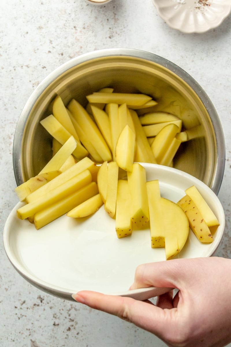 A hand is shown pouring a plate of potato sticks into a metal bowl.