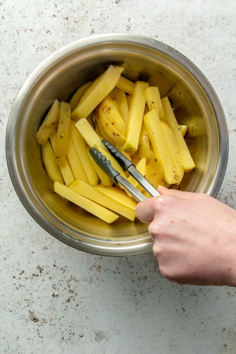 Sticks of potato sit in a metal mixing bowl. A hand is shown using tongs to mix the potatoes around.