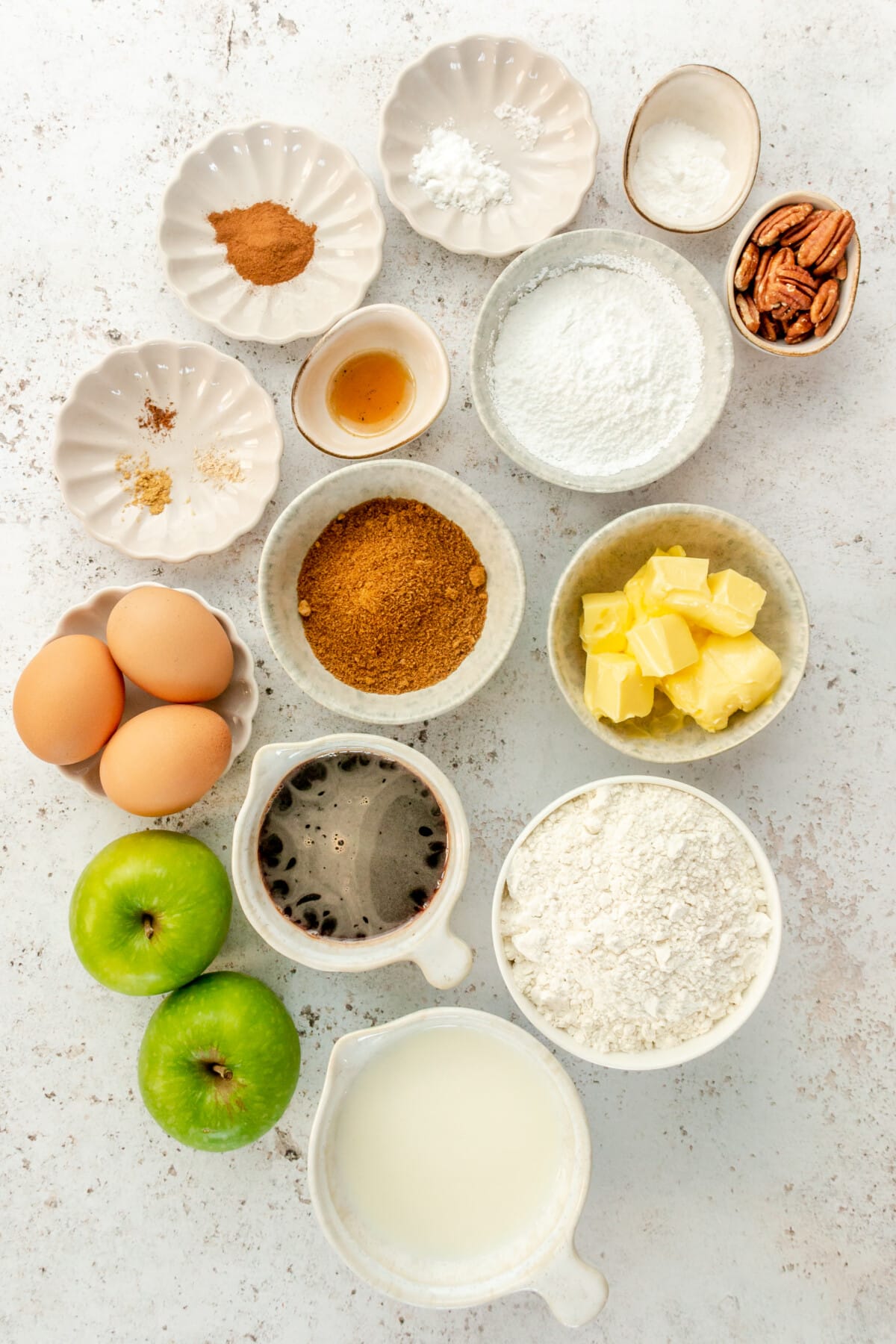 Ingredients for apple crumb cake sit in a variety of bowls and plates on a light grey surface.