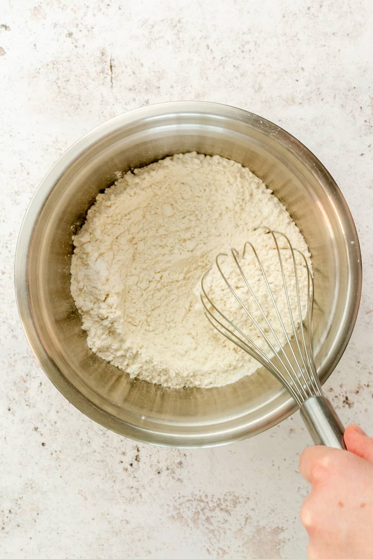 A flour mixture is whisked in a stainless steel bowl on a light grey surface.