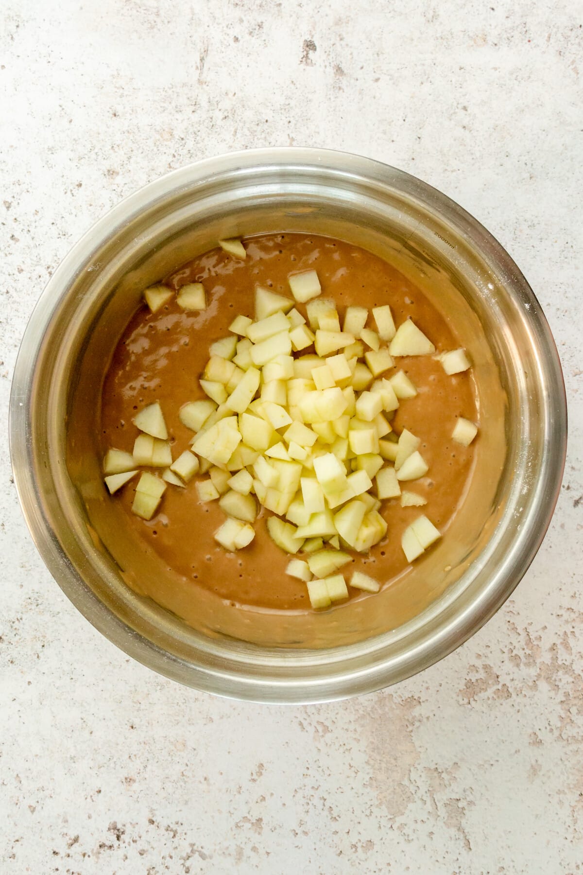 Cubes of apple sit in an apple crumb cake batter in a stainless steel bowl on a light grey surface.