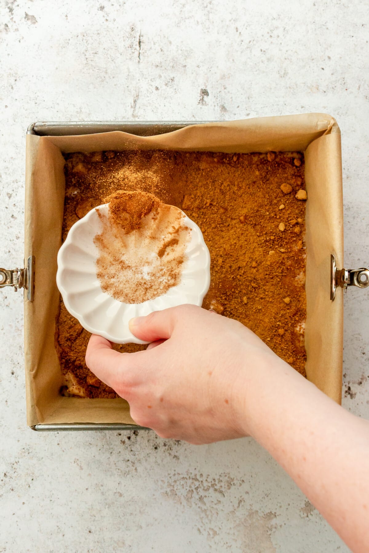 Ground cinnamon is tossed over an apple crumb cake batter in a square metal tin on a light grey surface.