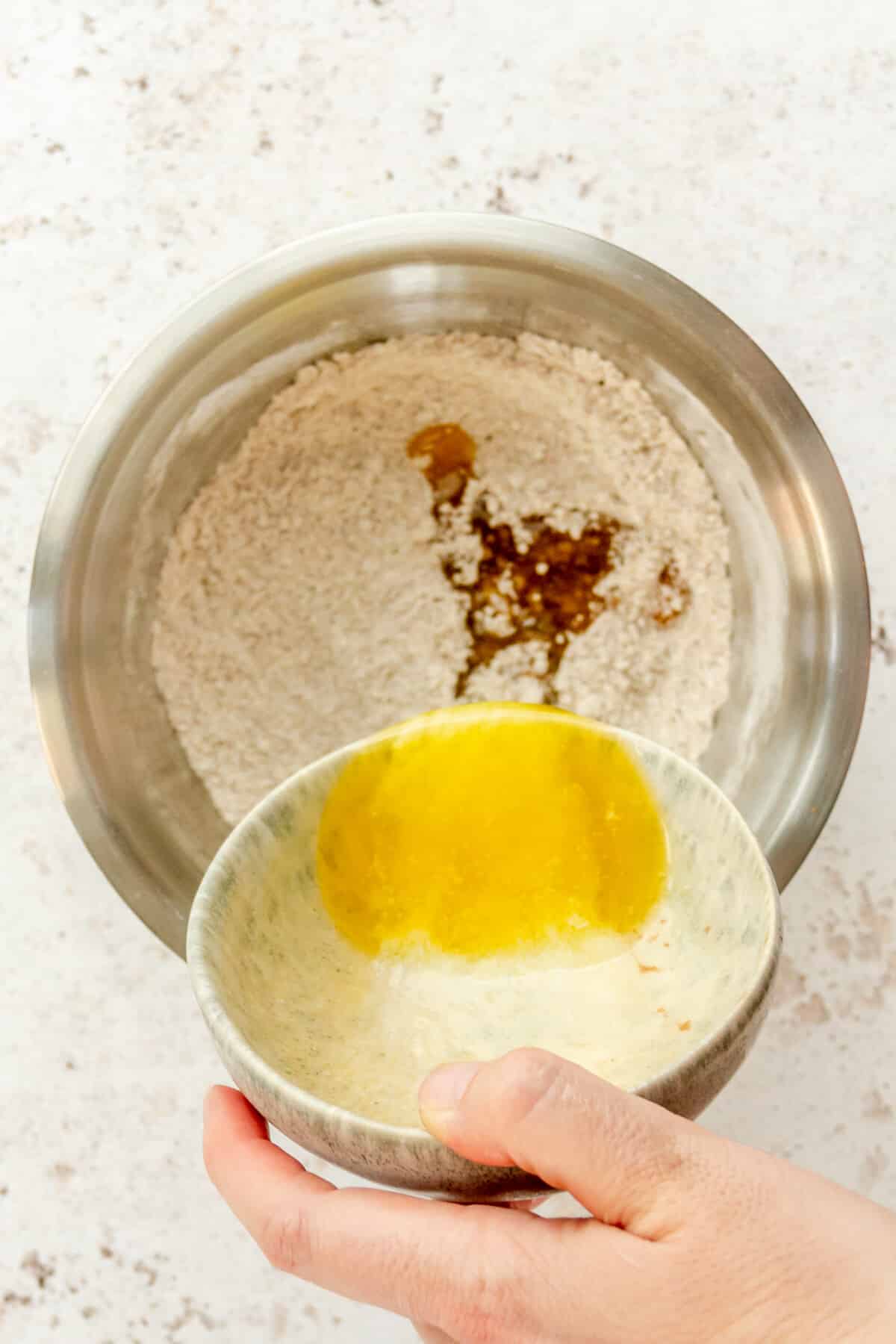 Melted butter is poured over a flour mixture in a stainless steel bowl on a light grey surface.