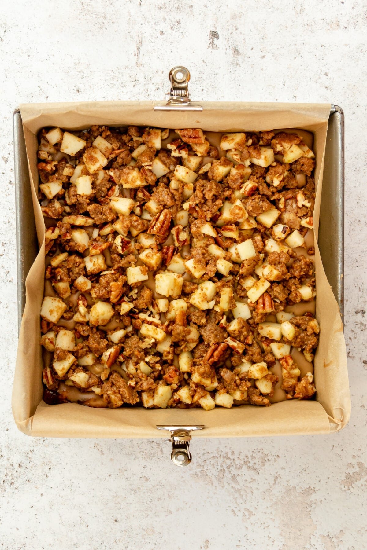 An apple crumb cake mixture sits in a square metal tin prior to baking on a light grey surface.