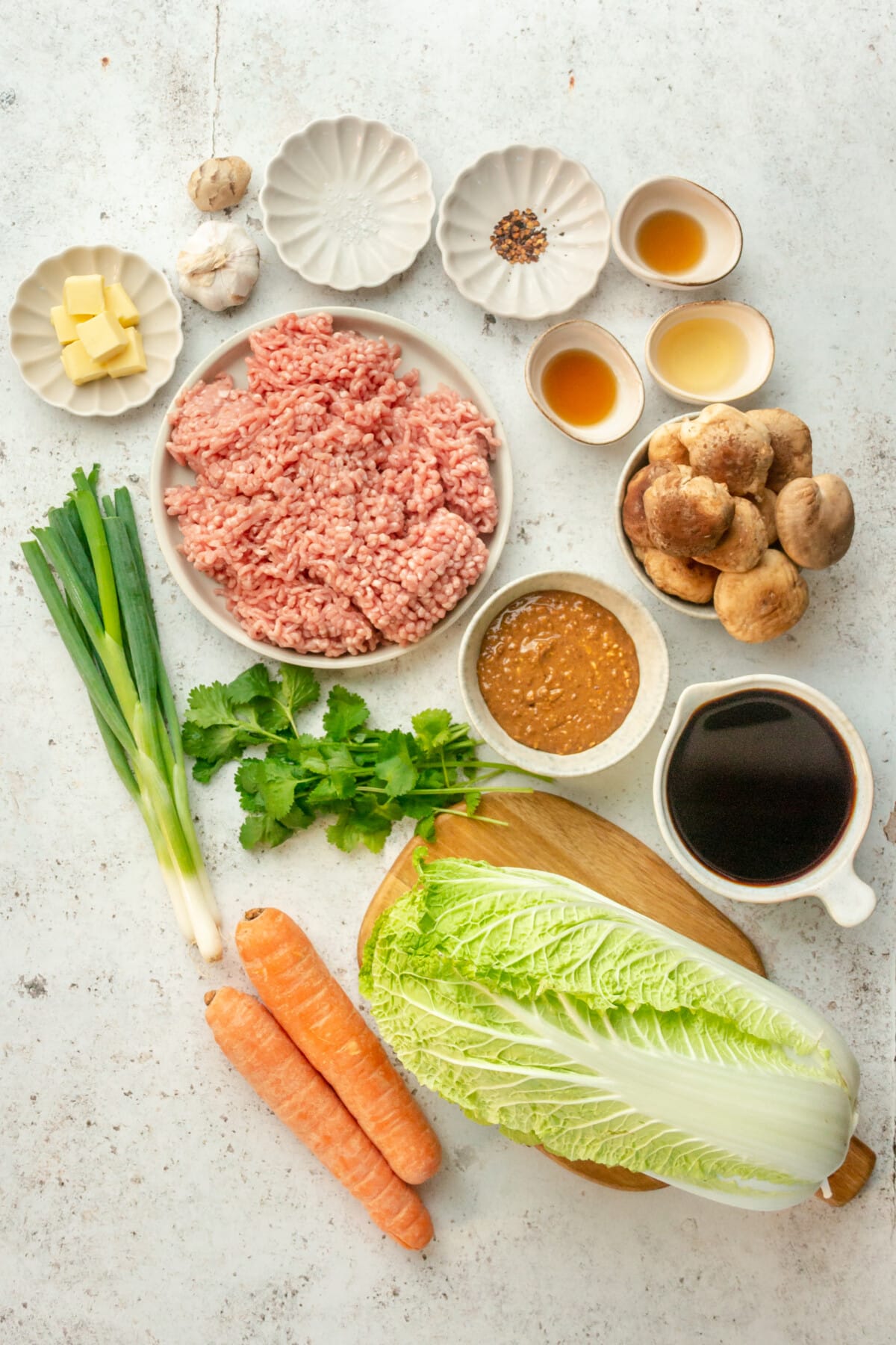 Ingredients for Asian style cabbage rolls with savory almond sauce sit in a variety of bowls and plates on a light grey surface.