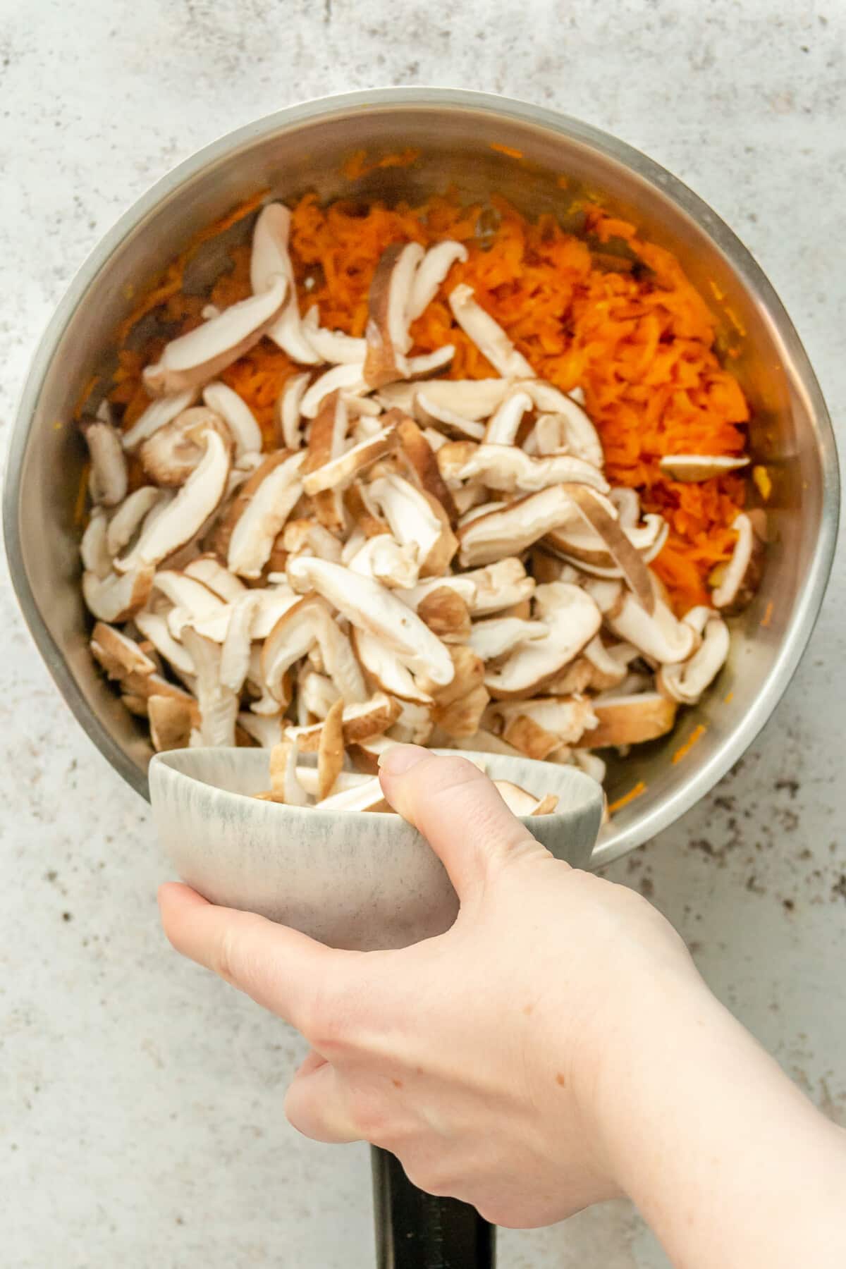 Sliced shiitake mushrooms are tossed over cooked shredded carrots in a stainless steel saucepan on a light gray surface.