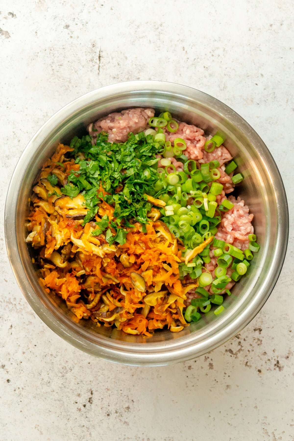 Cooked vegetables and chopped herbs and vegetables sit on top of pork mincemeat in a stainless steel bowl on a light gray surface.