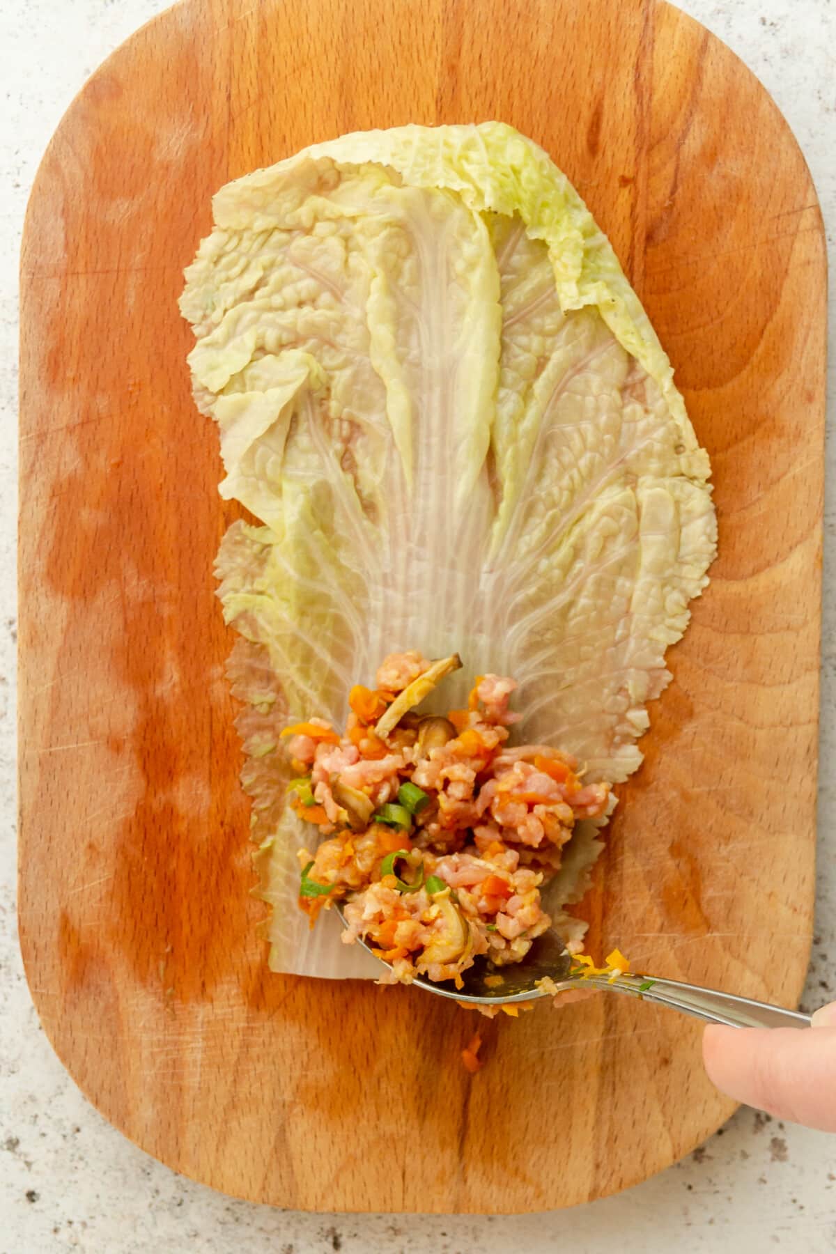 A pork and vegetable mincemeat filling is spooned onto the base of a napa cabbage leaf on a wooden board on a light gray surface.