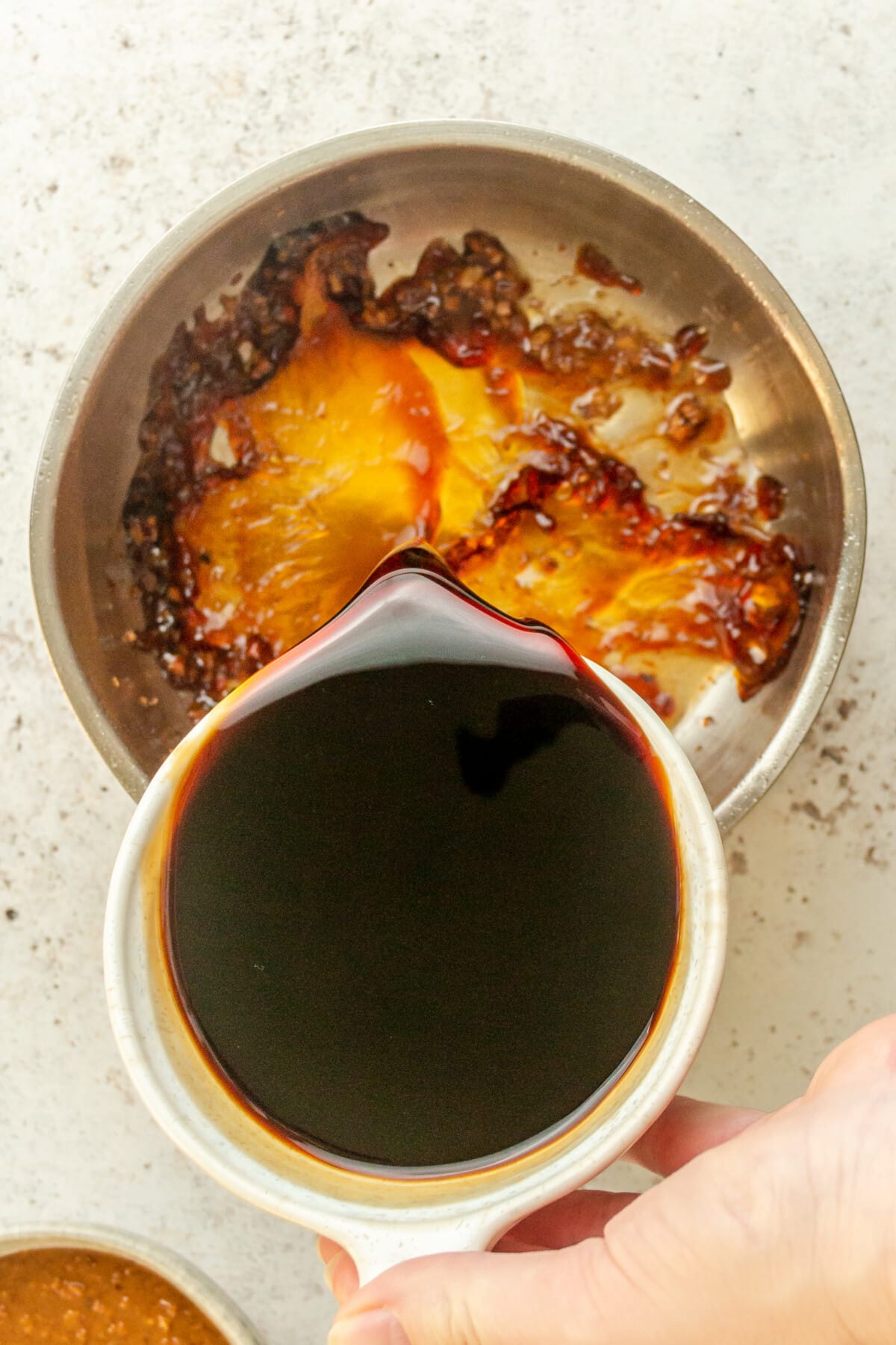 Coconut aminos is poured into a stainless steel saucepan on a light gray surface.