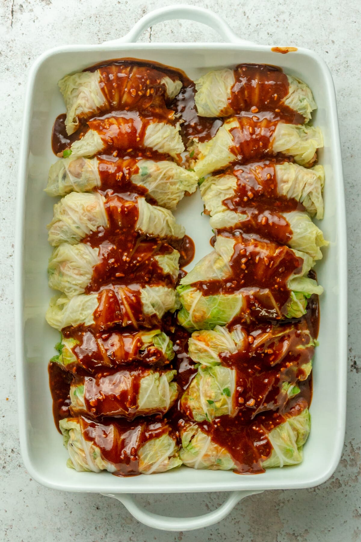 Asian style cabbage rolls sit in a rectangle white ceramic baking dish with a portion of savory almond sauce drizzled over on a light gray surface.