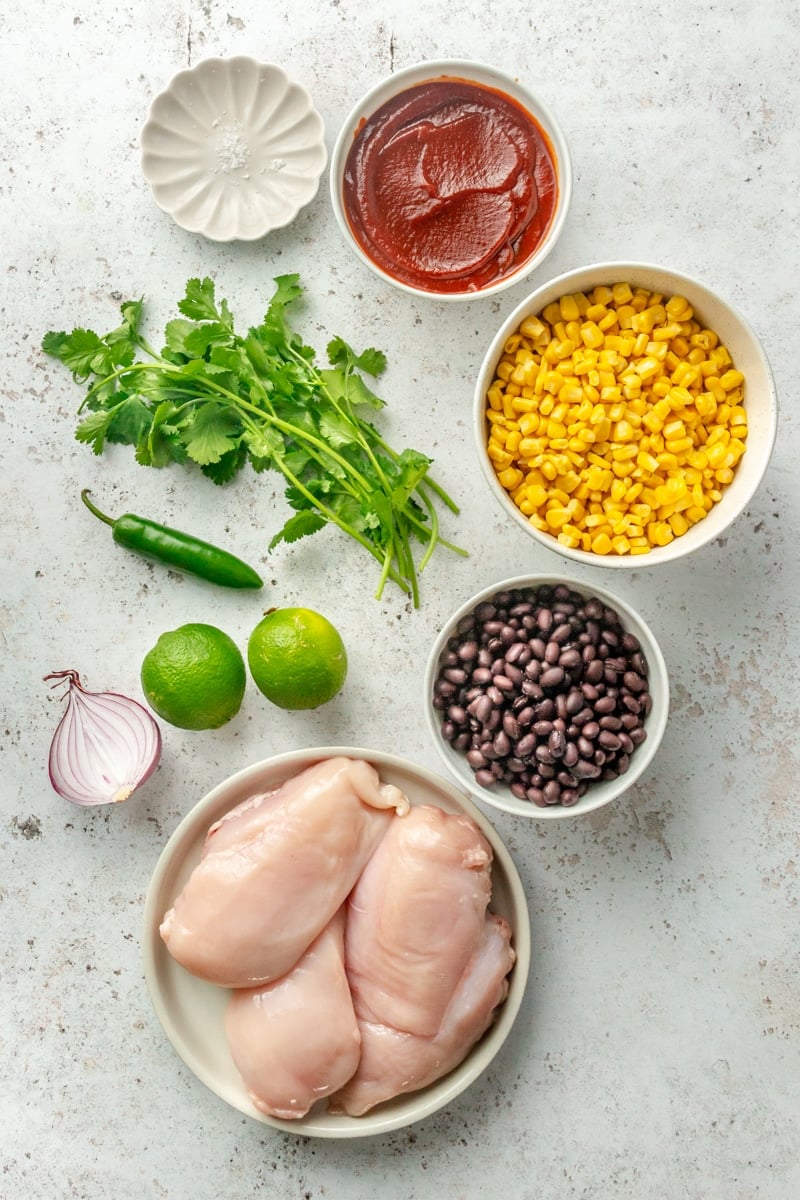 Ingredients for BBQ chicken tacos with black bean and corn salsa sit in a variety of bowls on a light grey colored surface.