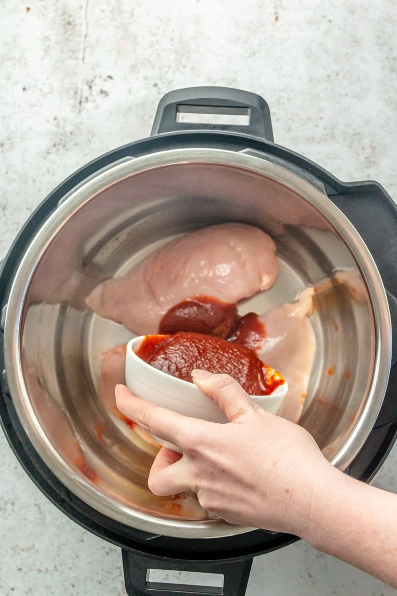 Three chicken breasts sit in the bottom of a pressure cooker. BBQ sauce is shown being poured over the top.